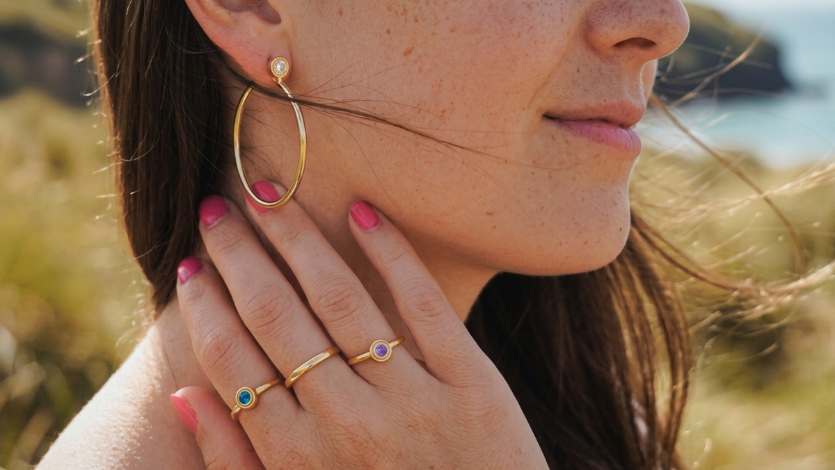 Woman wearing Elizabeth Paige gold hoop earrings and stacked IT Collection rings with colorful stones